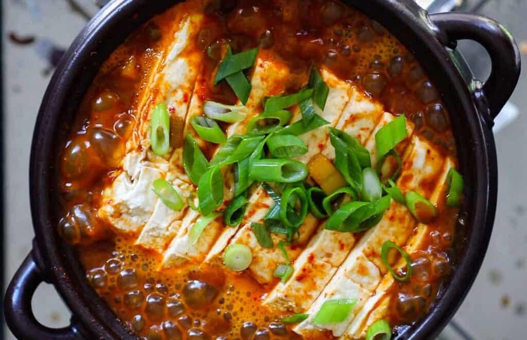 A bowl of bubbling doenjang jjigae is topped with sliced tofu and garnished with chopped green onions. The stew appears to have a rich, spicy red broth, served in a dark, circular pot.